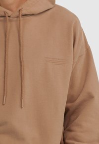 Felpa beige con cappuccio e lacci, con piccolo testo in rilievo "Young Poets Society" sul lato sinistro del petto, indossata da una persona mostrata dalla nuca fino a metà torso.