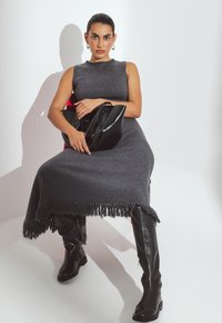 Naine, kellel on siledaks kammitud juuksed, kannab ilma varrukateta halli kleiti ja musti põlvini ulatuvaid saapaid, hoiab musta käekotti ning istub valge tausta ees.