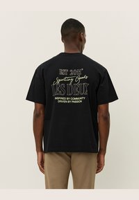 Tricou negru din bumbac cu maneci scurte, având un text grafic pe spate: "EST 2011 Sporting Goods LES DEUX Inspirat de Comunitate, Condus de Pasiune."