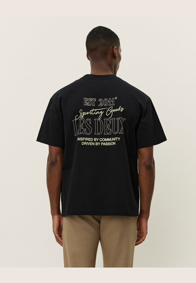 Tricou negru din bumbac cu maneci scurte, având un text grafic pe spate: "EST 2011 Sporting Goods LES DEUX Inspirat de Comunitate, Condus de Pasiune."