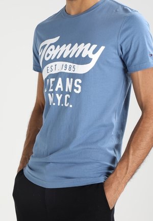 Mies sinisessä Tommy Jeans -T-paidassa, jossa valkoinen teksti "Tommy Est. 1985 Jeans N.Y.C.", ja mustissa housuissa, kädet taskuissa.