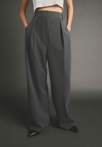 Pantaloni a vita alta e gamba larga in tessuto grigio scuro con finitura testurizzata, caratterizzati da pieghe frontali e tasche laterali. Scarpe nere a punta.