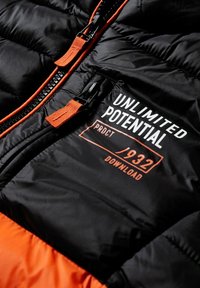 Czarna puchowa kurtka z pomarańczowymi akcentami, wyposażona w zamek błyskawiczny oraz napis "UNLIMITED POTENTIAL PRDCT 932 DOWNLOAD" w kolorze białym i pomarańczowym.