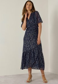 Abito navy con design avvolgente, maniche corte e orlo arricciato. Presenta motivi a pois bianchi su tutta la superficie. Abbinato a tacchi metallici con cinturini.