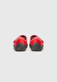 Chaussures en tissu rouge avec une texture lisse, dotées d'une sangle en velcro. La semelle est en caoutchouc noir, affichant un motif texturé pour une bonne adhérence.