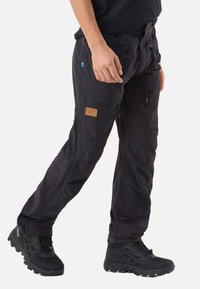 Čierne nákladné nohavice so zapínaním na zips na vreckách, hnedou koženou záplatou a detailmi na posilnenie kolien. Nosia sa s čiernymi outdoorovými topánkami.