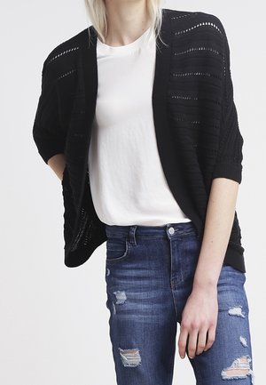 Frau trägt ein weißes T-Shirt, einen schwarzen offenen Strickcardigan mit halblangen Ärmeln und zerrissene blaue Jeans, steht vor hellem Hintergrund.