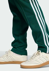 Zelené športové nohavice s bielymi trojitými pruhmi, spárované s smotanovými teniskami s textúrovanými akcentmi a gumenou podrážkou.