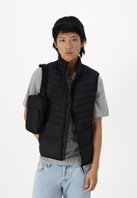 Zwart gewatteerd vest met verticale gestikt patroon, met rits aan de voorkant, gedragen over een grijze shirt. Kleine zwarte crossbody tas bevestigd aan de linkerkant.