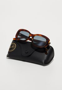 Ochelari de soare cu lentile gri, cu montură în carapace de țestoasă, așezați pe un capac dintr-o piele texturată neagră, având un logo aurit și sistem de închidere tip snap.