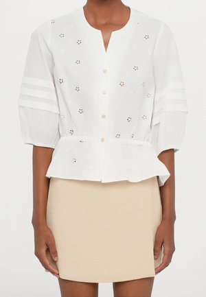 Biała bluzka na guziki z ażurowymi kwiatowymi detalami i rękawami o długości 3/4 z bufkami, zestawiona z beżową mini spódnicą.