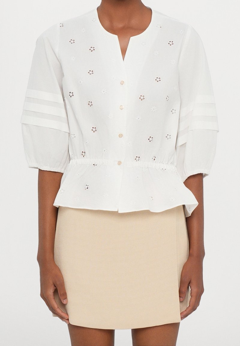 Blouse blanche boutonnée avec détails floraux en œillet et manches trois-quarts bouffantes, associée à une mini-jupe beige.