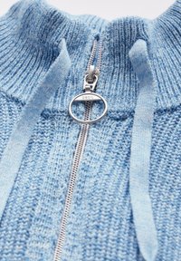 Lichtblauwe gebreide trui met een hoge kraag, zilveren rits aan de voorkant en een textuurpatroon door de stof heen.