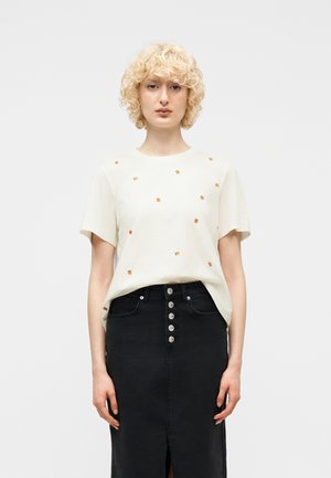 Kobieta o kręconych blond włosach nosi białą koszulę z małymi pomarańczowymi nadrukami oraz czarną spódnicę zapinaną na guziki z wysokim stanem i rozcięciem z przodu.