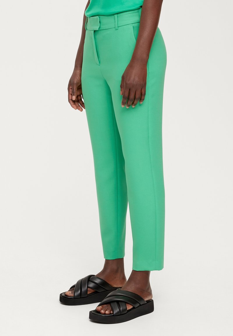 Jambes d'une personne portant un pantalon ajusté vert vif et des sandales plateformes noires avec de larges sangles croisées sur fond blanc.