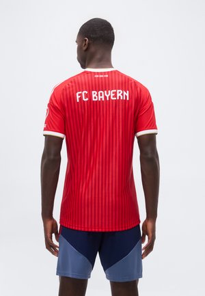 Mężczyzna ubrany w czerwoną koszulkę piłkarską FC Bayern z pionowymi paskami i granatowe krótkie spodenki, zwrócony tyłem na jednolitym tle.