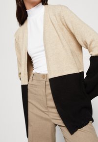 Kvinna som bär en beige och svart färgblockerande kofta, vit polotröja och beiga hög midja byxor, med handen i fickan och hoopörhängen.