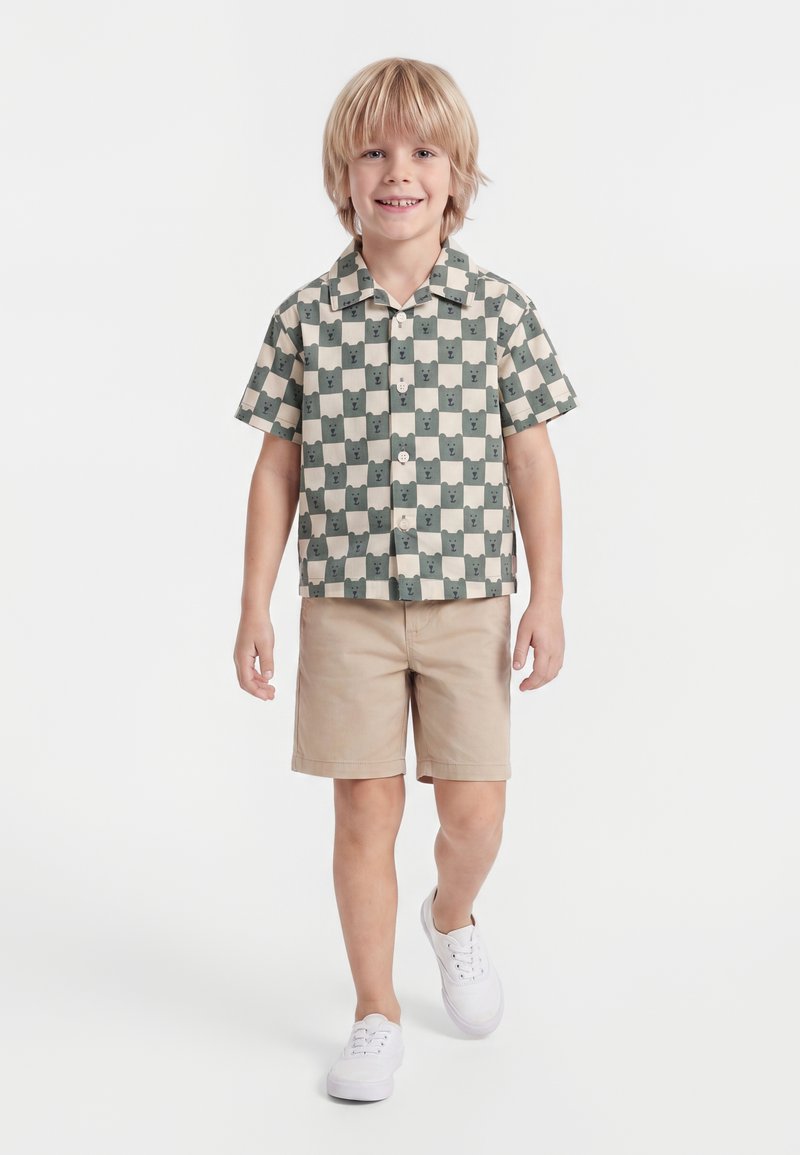 Jeune garçon marchant, portant une chemise à manches courtes à carreaux avec des visages d'ours, un short beige et des baskets blanches sur fond blanc.