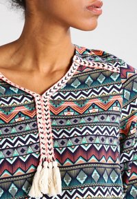 Mönstrad topp i flera färger med geometriska mönster i röd, blå och grön. V-ringning med flätade detaljer och fransar i mitten.
