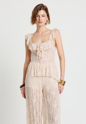 Femme portant un haut sans manches en dentelle rose pâle à volants et un pantalon large en dentelle assorti, accessoirisée avec des bracelets en or et en bois.