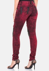 Personne debout, vêtue d’un jean skinny rouge effet délavé avec des détails de fermetures éclair noires, des sandales à talons hauts noires cloutées et un haut noir.
