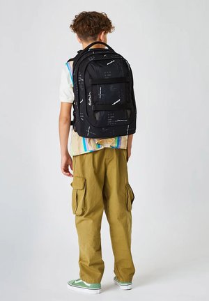 Kind steht mit dem Rücken zur Kamera, trägt einen schwarzen Rucksack mit Muster, ein bunt gestreiftes Hemd, khakifarbene Cargohosen und grüne Turnschuhe vor weißem Hintergrund.