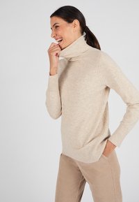 Beige coltrui met ribbelmanchetten en -zoom. Zachte textuur, gemiddelde dikte, met diagonale naden, gecombineerd met lichtbruine corduroy broek.