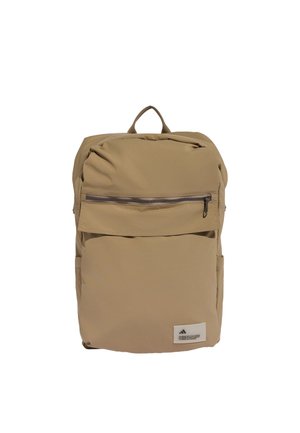 Mochila beige con asa superior, bolsillo frontal con cremallera y pequeña etiqueta blanca con texto y logo negro en la parte inferior frontal.