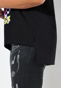 Sort bomulds tshirt med et grafisk print, der har farverige tekster, sideslids og en afslappet pasform, parret med slidte mørke jeans.