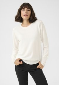 Weißer Langarm-Pullover aus weichem Stoff mit rundem Halsausschnitt und gerippten Bündchen, kombiniert mit schwarzen, figurbetonten Hosen.
