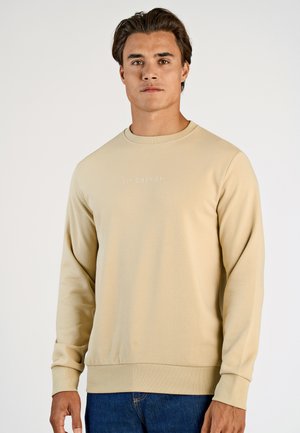 Beige sweatshirt med lange ærmer og rund hals, med en subtil logo-tekst i lys farve på forsiden. Blød stofstruktur.