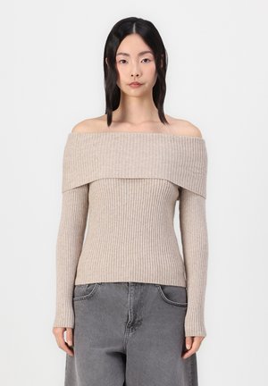 Młoda kobieta z prostymi czarnymi włosami, ubrana w beżowy sweter na jedno ramię z prążkowanej dzianiny oraz luźne szare dżinsy, stojąca na tle jednolitego białego tła.