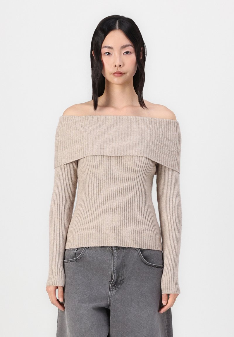 Giovane donna con capelli neri lisci che indossa un maglione beige a coste, scollato sulle spalle, e jeans grigi larghi, in piedi contro uno sfondo bianco semplice.