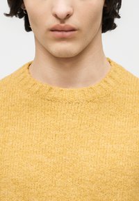 Gros plan sur une personne portant un pull en maille moutarde douce et texturée, avec un col rond et une petite boucle d'oreille.