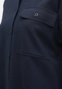 Manteau bleu marine foncé avec boutons sur le devant et une grande poche à rabat boutonnée sur la poitrine.