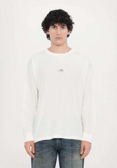 Camiseta blanca de manga larga con cuello redondo y un pequeño gráfico rectangular en el pecho. Tejido suave, corte holgado.