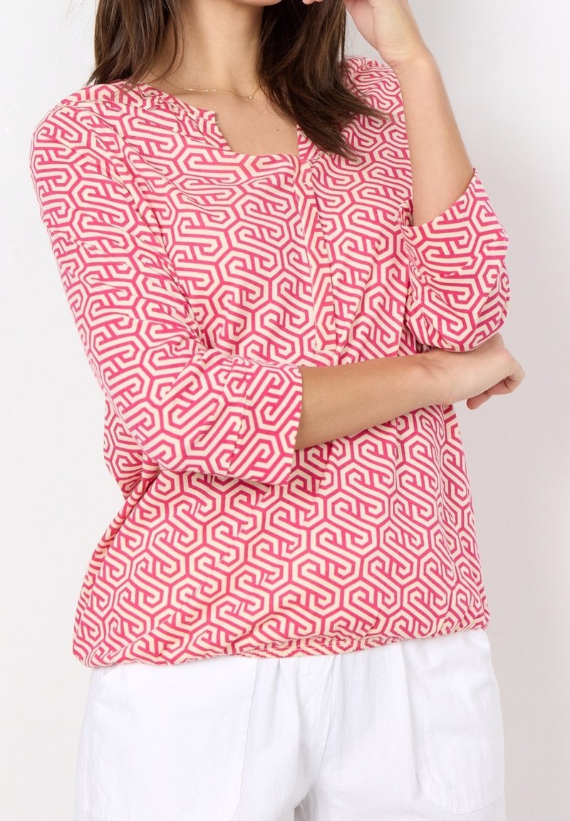 Femme portant un chemisier à motifs géométriques rose et blanc avec des manches trois-quarts et un pantalon blanc, posant avec un bras croisé.
