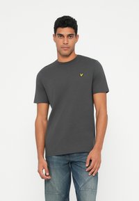 Jovem vestindo uma t-shirt de manga curta cinza escuro com um pequeno logotipo de um pássaro amarelo no peito e jeans azul claro contra um fundo branco.