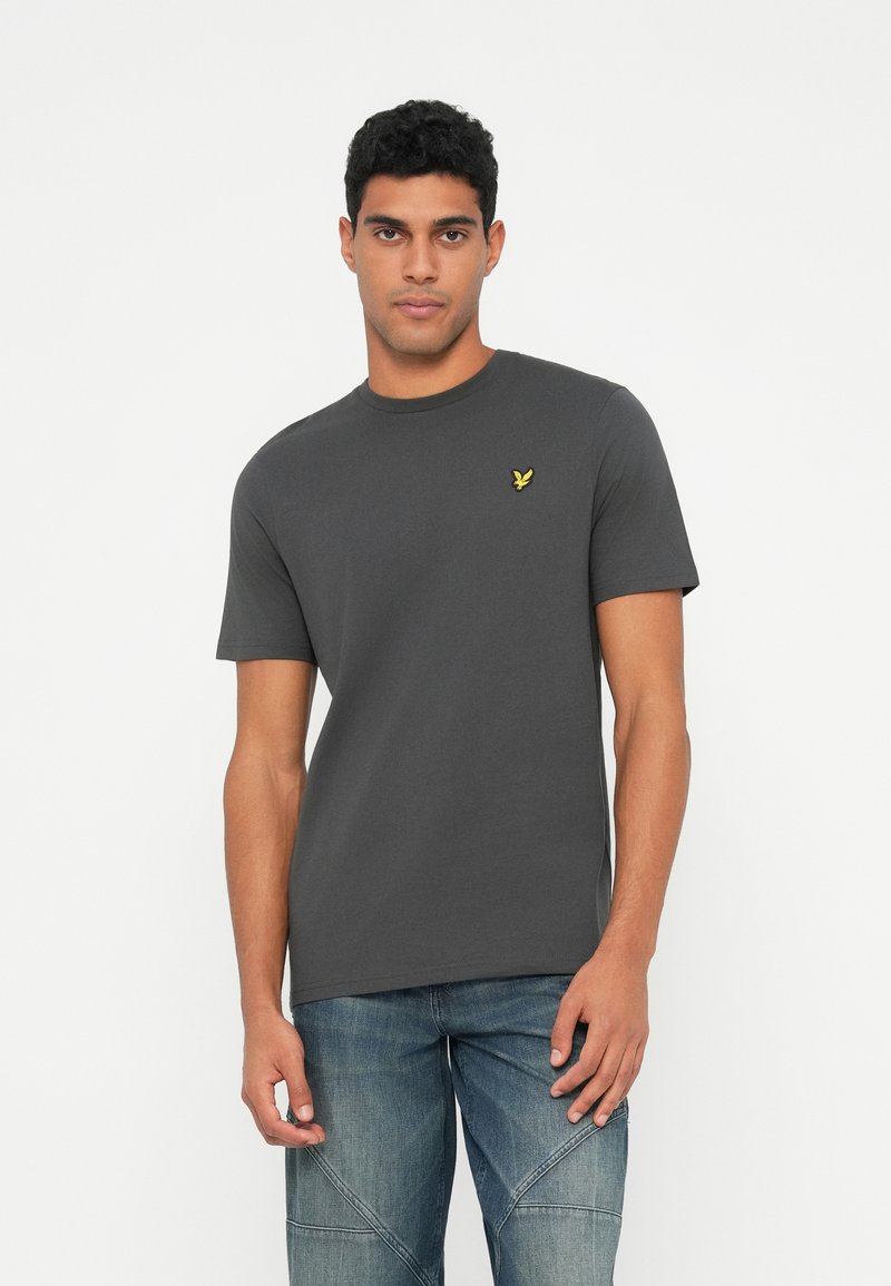 Jovem vestindo uma t-shirt de manga curta cinza escuro com um pequeno logotipo de um pássaro amarelo no peito e jeans azul claro contra um fundo branco.