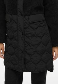 Melnās mētelis ar kviltētu apakšdaļu un pūkainu augšdaļu, ar drukājamām pogām un kabatām praktiskai lietošanai. Mīksta tekstūra visā virsmas garumā.