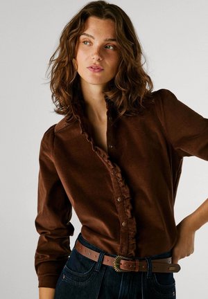 Brun fløjlsskjorte med rynket front og knaplukning, parret med mørke denim jeans og et brunt bælte med en rund spænde.