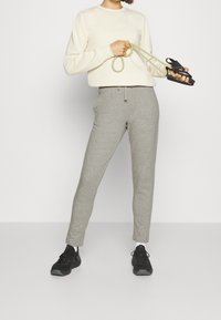 Light cream sweatshirt, gray tapered sweatpants, and black athletic sneakers. Model holds a green resistance band with black handles.