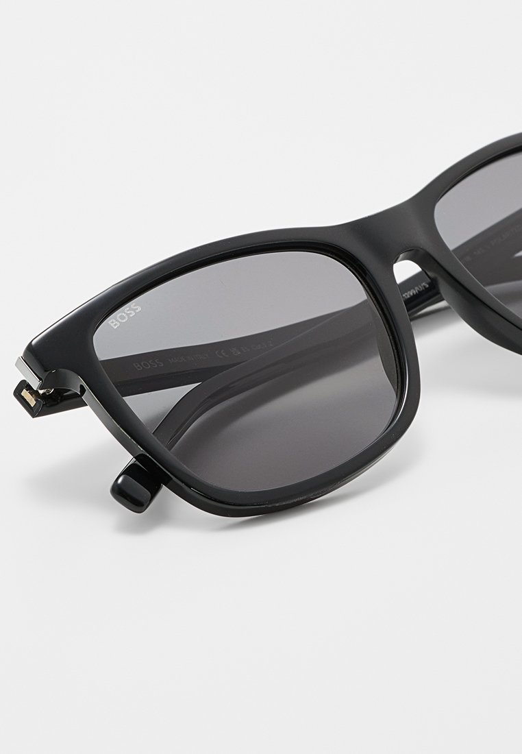 Schwarze rechteckige Sonnenbrille mit grauen Gläsern. Besitzt eine glänzende Oberfläche, einen stabilen Rahmen und metallic Akzente in der Nähe der Scharniere.