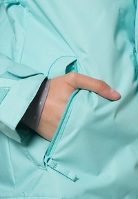 Veste turquoise avec une finition mate et lisse. Comprend une poche latérale avec une fermeture éclair turquoise et une patte de fermeture sur les manches.