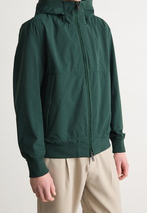 Man wearing a dark green hooded zip-up jacket with ribbed cuffs and beige pants, standing against a plain background.