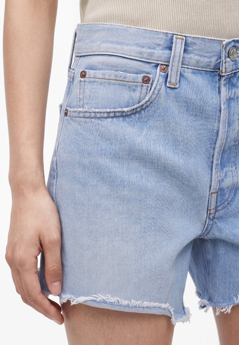 Short en denim bleu clair avec des ourlets effilochés porté par une personne, montrant les détails des poches avant et une partie de sa main reposant à proximité.