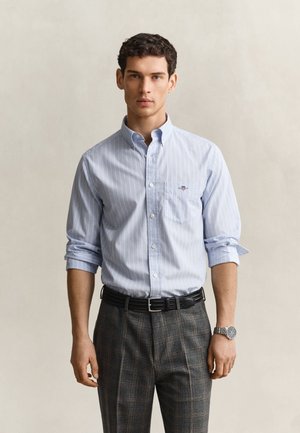 Jeune homme portant une chemise à rayures bleu clair avec les manches retroussées et un pantalon gris à carreaux avec une ceinture noire et une montre-bracelet.