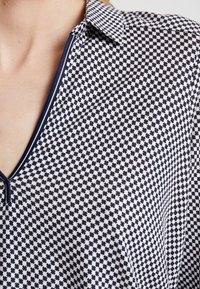 Top a righe blu navy e bianco con scollatura a V, colletto e piccoli motivi geometrici su tutta la superficie. Tessuto morbido e strutturato.