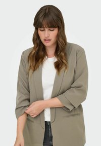 Blazer verde claro con un diseño a medida, mangas de tres cuartos, bolsillos laterales y una textura suave, llevado sobre una camiseta blanca.