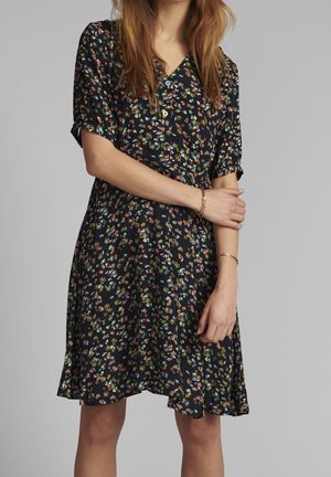 Kobieta w czarnej sukience w kwiaty z wielobarwnymi, małymi kwiatami, z krótkimi rękawami, zapinana z przodu na guziki, z złotymi bransoletkami na obu nadgarstkach.